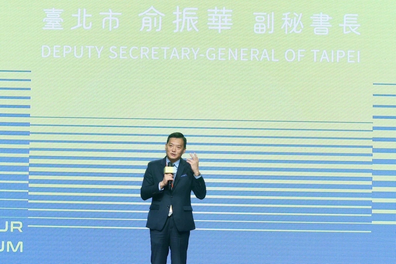 臺北市副秘書長俞振華於活動舞台上發表演說，背景螢幕顯示其職稱英文為 Deputy Secretary-General of Taipei，畫面以藍綠色線條圖形構成。