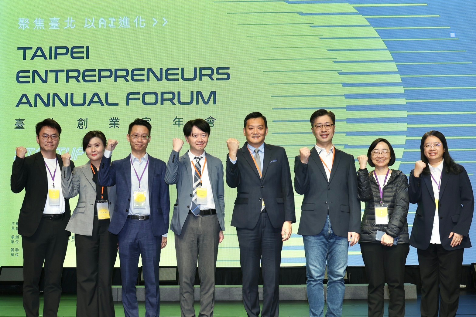 八位出席貴賓站立於舞台前合影，手握拳展現團結氣勢，背景為臺北創業家年會主視覺螢幕，顯示活動名稱「TAIPEI ENTREPRENEURS ANNUAL FORUM」與「聚焦臺北 以創式進化」標語。