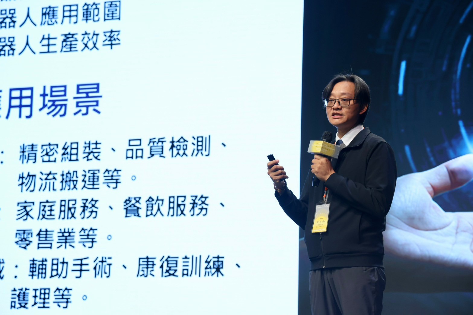 一位男講者身穿黑色外套與眼鏡，手持簡報筆與麥克風，在螢幕前講解機器人應用場景，背景文字顯示「精密組裝、品質檢測、物流搬運、家庭服務、餐飲服務、零售業、輔助手術、康復訓練、護理等」。