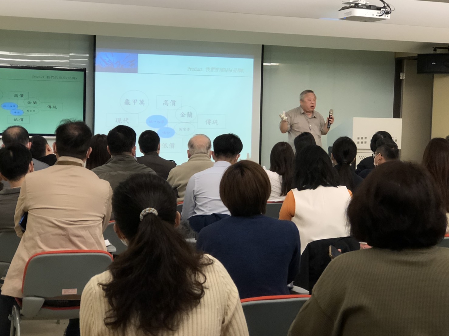 一位資深講者，黃禾國際廣告有限公司營運董事，何清輝，於教室內以投影簡報講解市場定位與產品價值，手持麥克風與教具，面對數十位成人學員，學員專注聆聽，呈現企業培訓或成人教育的專業學習場景。